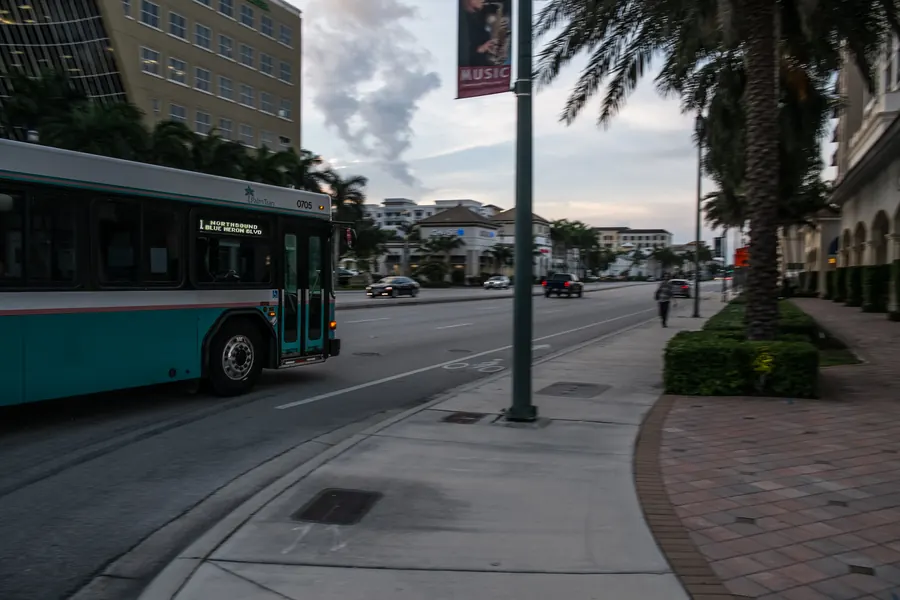 A city bus passing palms and a brick sidewalk on a downtown Boca Raton street