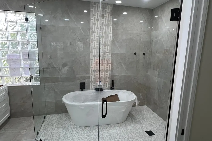 A frameless glass enclosure around a freestanding tub and shower, with matte black clamps and a black door pull, gray marble-look walls and a white pebble mosaic floor