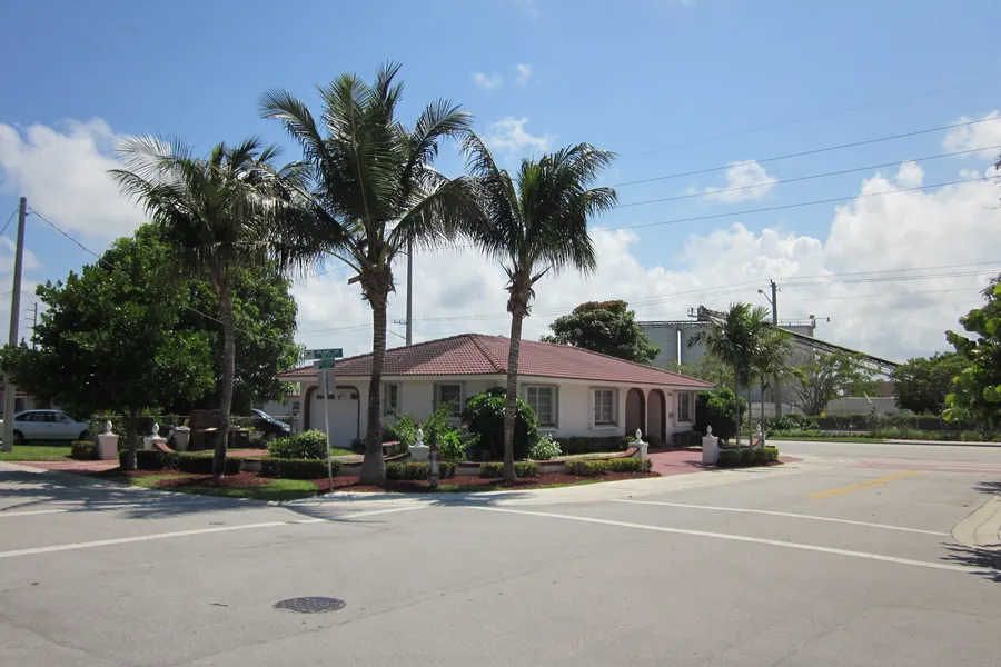 A single-story house with a red tile roof behind three tall palms, on a street corner in Pearl City, Boca Raton