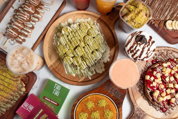 An overhead spread of Tiki's desserts and drinks: knafa, a waffle stick, crêpes, a shake and iced coffee on wooden boards