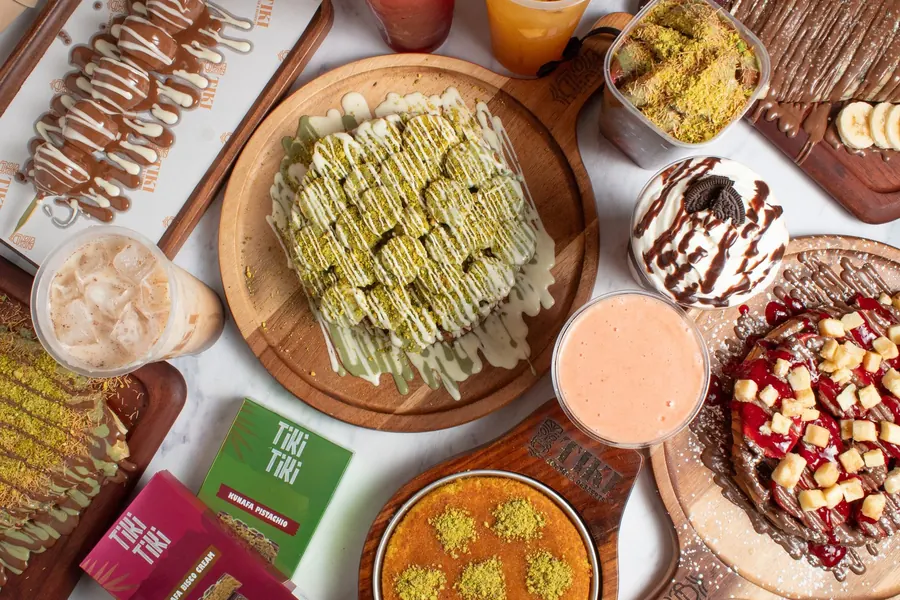 An overhead spread of Tiki's desserts and drinks: knafa, a waffle stick, crêpes, a shake and iced coffee on wooden boards